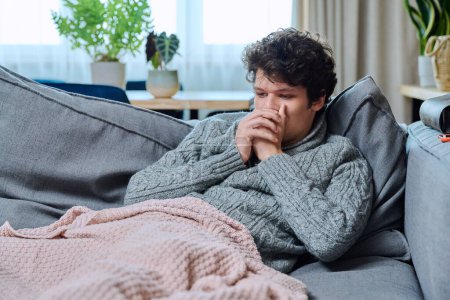 Foto de Joven enfermo tosiendo, con signos de enfermedad infecciosa respiratoria, en suéter de lana sentado en el sofá en casa. Goteo nasal, tos, estación fría - Imagen libre de derechos