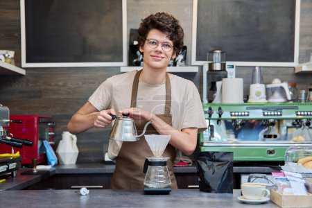 Portrait of young guy barista at counter in coffee shop. Handsome curly-haired male worker preparing coffee drinks looking at camera. Food service occupation, small business, work, employee, staff