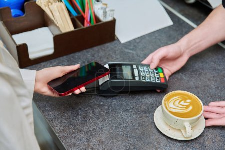 Foto de Primer plano de una mano de los clientes con un teléfono inteligente que paga por una taza de café en un café, sin contacto con el uso de la terminal, pago móvil - Imagen libre de derechos