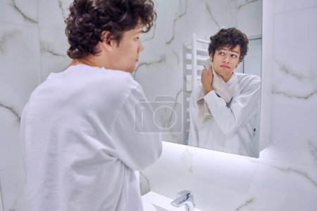 Téléchargez les photos : Beau jeune homme 18-20 ans dans la salle de bain avec serviette regardant son reflet dans le miroir. Matin soir routine, hygiène, soins de la peau cosmétiques pour le visage pour les jeunes hommes - en image libre de droit