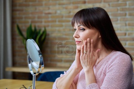 Middle aged beautiful woman looking at face in mirror. Professional medical care cosmetics, use of spa massage, beauty injections. Skincare, plastic surgery, age beauty skin care, cosmetology concept