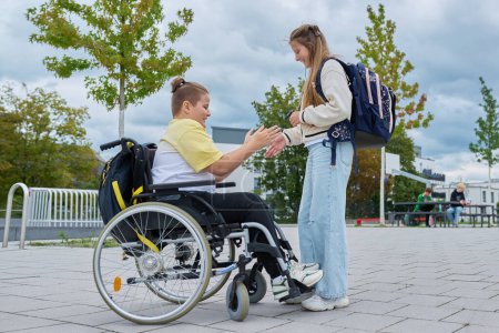 Children 10, 11 years old friends classmates disabled boy in wheelchair and girl with backpack talking having fun near school building. Education, friendship, communication, disability, inclusiveness