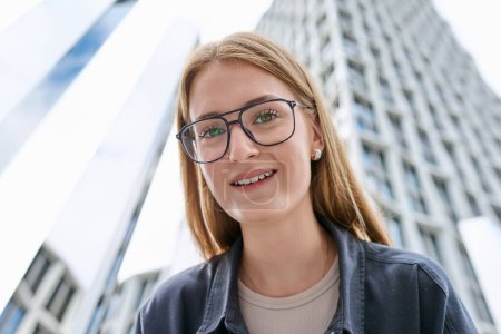 Photo for Closeup face, teenage beautiful female in glasses looking at camera, modern glass skyscrapers, urban style background. Youth, lifestyle, city life, young people concept - Royalty Free Image
