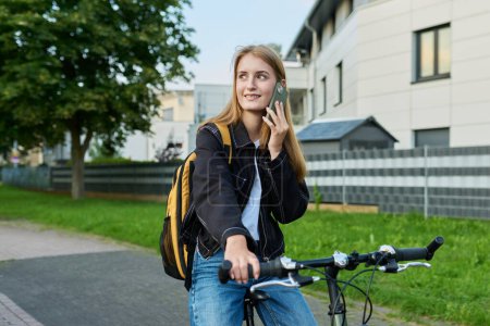Téléchargez les photos : Portrait d'une étudiante adolescente avec sac à dos parlant au téléphone à vélo. Souriant adolescent femme 16, 17 ans en plein air sur la route. École secondaire, mode de vie, adolescence, concept jeunesse - en image libre de droit
