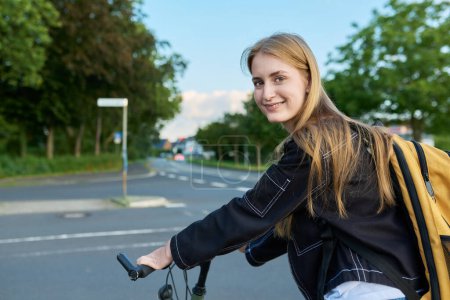 Téléchargez les photos : Portrait d'une étudiante adolescente avec sac à dos à vélo. Souriant adolescent femme 16, 17 ans regardant la caméra à l'extérieur sur la route. École secondaire, mode de vie, adolescence, concept jeunesse - en image libre de droit