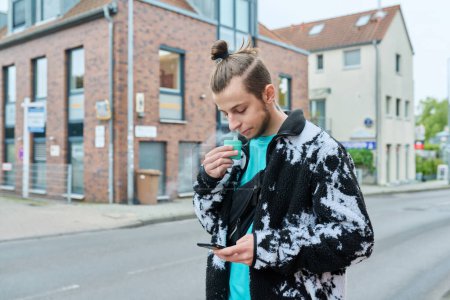Foto de Joven hipster masculino con peinado de barba de moda usando smartphone, fumar cigarrillo electrónico desechable, al aire libre en la calle de la ciudad. Juventud, estilo de vida, malos hábitos, tecnología, aplicaciones móviles - Imagen libre de derechos