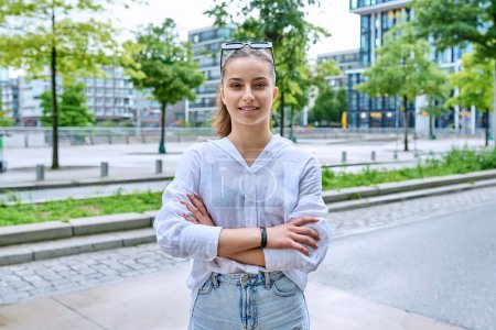 Téléchargez les photos : Portrait d'adolescente souriante confiante regardant la caméra, avec les bras croisés sur la rue de la ville moderne. Adolescence, mode de vie urbain, concept des jeunes - en image libre de droit
