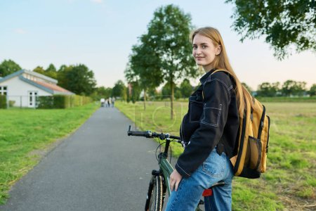 Téléchargez les photos : Portrait d'une étudiante adolescente avec sac à dos à vélo. Souriant adolescent femme 16, 17 ans regardant la caméra à l'extérieur sur la route, copier l'espace. École secondaire, mode de vie, adolescence, concept jeunesse - en image libre de droit