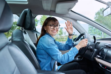 Téléchargez les photos : Femme d'âge moyen conducteur portant des lunettes assis au volant dans la voiture, souriant en regardant la caméra. Transport, mode de vie, conduite, achat assurance réparation entretien de voiture, concept de personnes matures - en image libre de droit