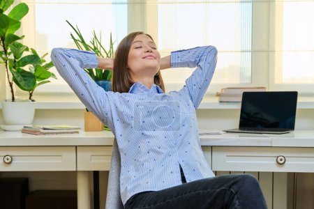 Entspannte junge Frau mit geschlossenen Augen und den Händen hinter dem Kopf, die auf einem Stuhl neben einem Tisch mit Computer-Laptop sitzt. Ruhige Studentin nach Online-Unterricht, junge Freiberuflerin, die aus der Ferne arbeitet