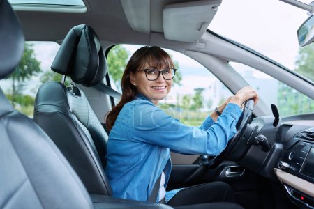 Téléchargez les photos : Femme d'âge moyen conducteur portant des lunettes assis au volant dans la voiture, souriant en regardant la caméra. Transport, mode de vie, conduite, achat assurance réparation entretien de voiture, concept de personnes matures - en image libre de droit
