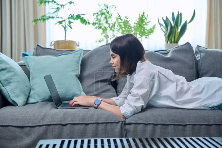 Photo for Young woman typing on laptop while lying on sofa at home. Serious female using computer for leisure work study communication, social media, blog, freelancing, internet technology, people concept - Royalty Free Image