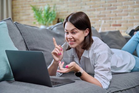 Freudig überrascht blickt eine junge Frau auf den Laptop-Bildschirm, der zu Hause auf dem Sofa liegt. Emotionale weibliche Nutzung des Computers in der Freizeit Studienkommunikation, Social Media Blog, Freelancing, Internet, Technologie