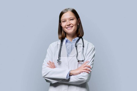 Téléchargez les photos : Portrait de jeune femme médecin infirmière étudiante en médecine stagiaire en manteau blanc sur fond de studio gris. Femme confiante avec les bras croisés regardant la caméra. Médecine, personnel médical professionnel - en image libre de droit