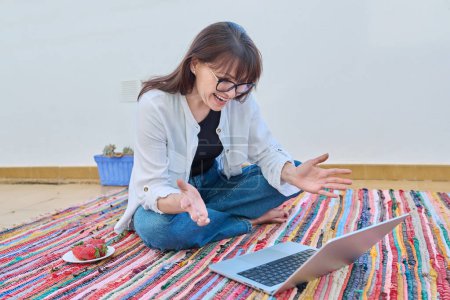 Photo for Happy middle-aged woman sitting on floor at home using laptop computer, with bowl of fresh strawberries. Leisure, work, communication, social media, blog, freelancing, internet technology, 40s people - Royalty Free Image