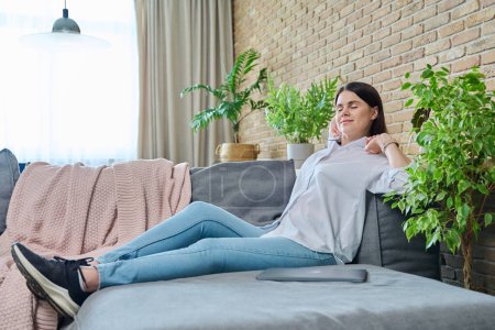 Relaxed young woman with raised arms sitting on sofa in living room. Calm happy female with closed eyes, resting, stretching back body, pleasant tired