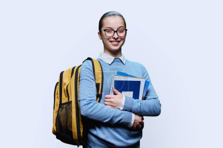 Téléchargez les photos : Portrait d'une adolescente lycéenne sur fond de studio blanc. Femme adolescente souriante dans des lunettes avec un manuel de sac à dos regardant la caméra. Éducation adolescence connaissance apprentissage mode de vie - en image libre de droit