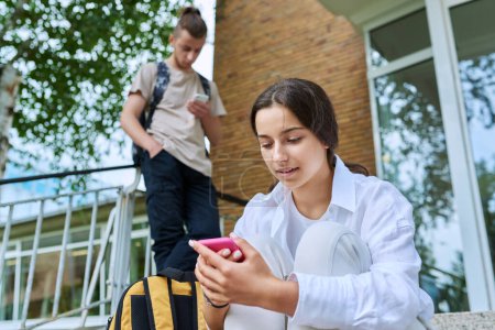 Téléchargez les photos : Les élèves adolescents avec des smartphones sur les étapes du bâtiment éducatif, en particulier les adolescentes d'âge secondaire en utilisant gadget. Adolescence, jeunesse, éducation, mode de vie, concept technologique - en image libre de droit