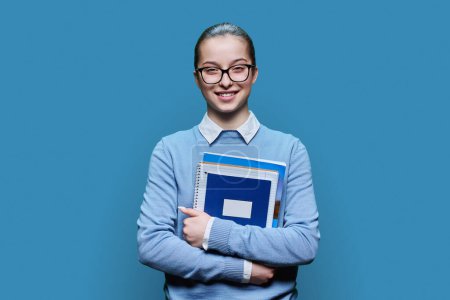 Téléchargez les photos : Portrait d'adolescente lycéenne sur fond de studio bleu. Adolescente souriante dans des lunettes avec des cahiers de manuels regardant la caméra. Éducation adolescence connaissance apprentissage mode de vie - en image libre de droit