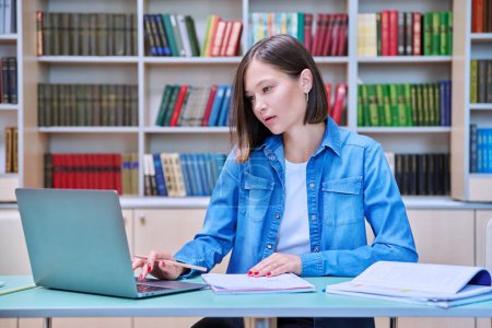 Photo for Young female university student studying inside library, typing on laptop computer. Knowledge, education, youth, college university concept - Royalty Free Image