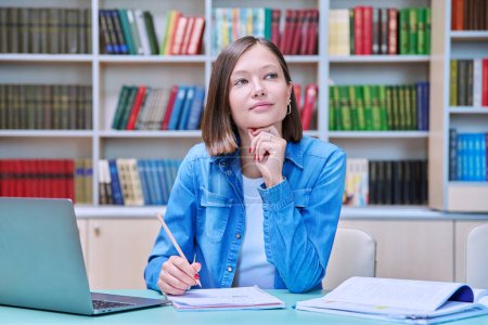 Photo for Portrait of smiling young female university student sitting at desk with laptop in library classroom of educational building. Knowledge, higher education, youth concept - Royalty Free Image