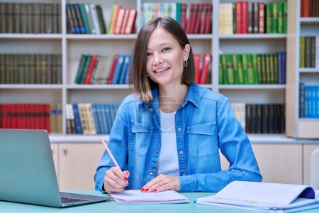 Photo for Portrait of smiling young female university student looking at camera sitting at desk with laptop in library classroom of educational building. Knowledge, higher education, youth concept - Royalty Free Image