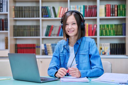 Photo for Portrait of smiling young female university student wearing headphones looking at camera sitting at desk with laptop in library classroom of educational building. Knowledge, education, youth concept - Royalty Free Image