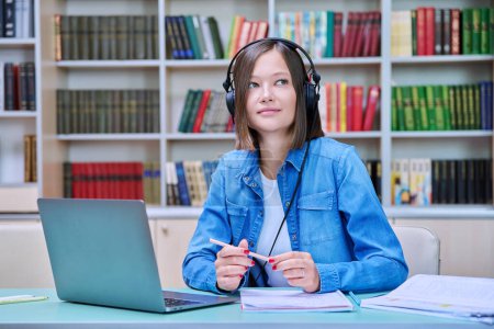 Photo for Portrait of smiling young female university student wearing headphones sitting at desk with laptop in library classroom of educational building. Knowledge, education, youth concept - Royalty Free Image