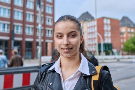 Téléchargez les photos : Portrait d'un lycéen adolescent, souriant fille confiante 16, 17 ans avec sac à dos regardant la caméra en plein air, dans la rue de la ville moderne. Vie urbaine, adolescence, éducation, concept de jeunesse - en image libre de droit