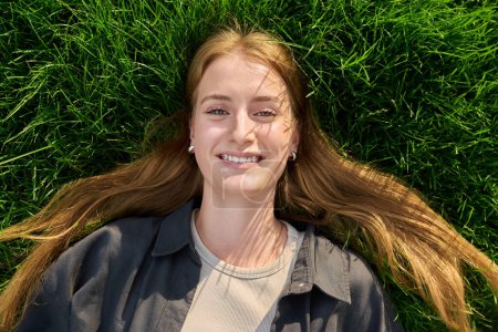 Photo for Top view, close-up of face of smiling blonde teenage girl lying on grass. Beautiful positive female with long hair looking at camera. Adolescence, youth, beauty, lifestyle concept - Royalty Free Image
