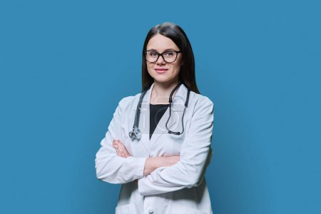 Photo for Portrait of friendly confident female doctor in white coat on blue studio background. Smiling medic with crossed arms looking at camera. Medicine, medical staff, treatment, health care concept - Royalty Free Image