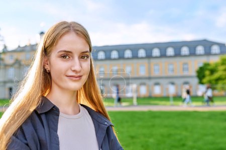 Photo for Headshot portrait of young teenage smiling female looking at camera outdoor, copy space green lawn grass educational building. Confident friendly cool girl in sunny sunset light. Youth, lifestyle - Royalty Free Image