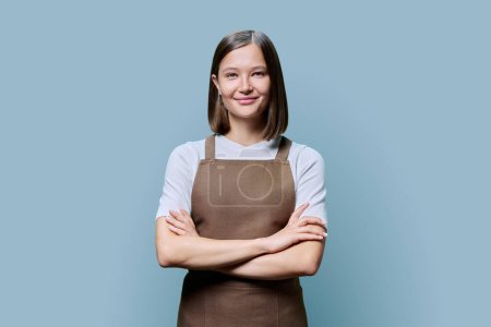 Photo for Young smiling woman in apron looking at camera on blue studio background. Confident female small business owner, service worker posing with arms crossed. Business, work, occupation, staff concept - Royalty Free Image
