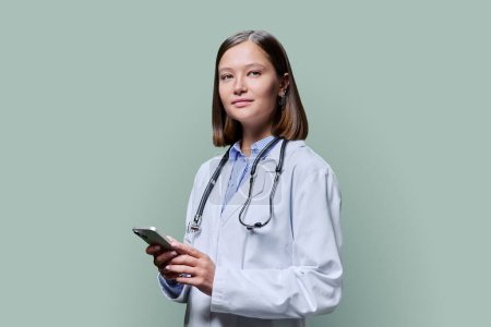 Photo for Young female doctor using smartphone on green background. Smiling friendly female in white coat with stethoscope looking at camera, technology for work, internet online medical applications, service - Royalty Free Image