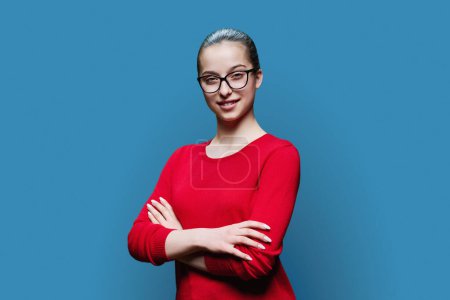 Téléchargez les photos : Portrait adolescent souriant confiant avec des lunettes en rouge sur fond bleu. Femme positive 16, 17 ans regardant la caméra avec les bras croisés lycéen style de vie adolescent adolescent - en image libre de droit