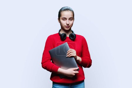 Téléchargez les photos : Portrait d'une adolescente étudiante dans un casque avec ordinateur portable dans les mains sur fond de studio blanc. Adolescente souriante en rouge regardant la caméra. Adolescence, éducation loisirs mode de vie technologie - en image libre de droit