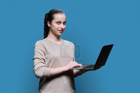 Téléchargez les photos : Adolescente lycéenne utilisant un ordinateur portable regardant la caméra sur fond de studio bleu. Technologie, e-learning, éducation, adolescence, concept jeunesse. - en image libre de droit