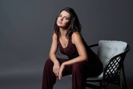 Téléchargez les photos : Jeune femme séduisante sensuelle à la mode posant assise sur une chaise sur fond de studio gris foncé. Beauté, mode, concept de style - en image libre de droit