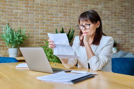 Photo for Serious mature businesswoman reading important official letter sitting at desk in office. Business work banking services taxes, contract statement agreement investment deal, paperwork correspondence - Royalty Free Image