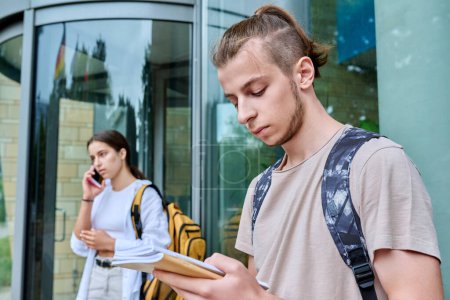 Foto de Hipster chico adolescente estudiante 18, 19 años de edad con mochila cuadernos de pie al aire libre, fondo del edificio educativo. Juventud, educación, concepto de estilo de vida - Imagen libre de derechos