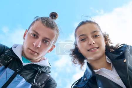 Photo for Closeup faces of young teenage guy and girl smiling looking down at camera, blue sky with clouds background. Youth communication leisure friendship lifestyle, college high school students education - Royalty Free Image