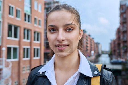 Téléchargez les photos : Portrait d'un lycéen adolescent, souriant fille confiante 16, 17 ans avec sac à dos regardant la caméra en plein air, dans la rue de la ville moderne. Vie urbaine, adolescence, éducation, concept de jeunesse - en image libre de droit