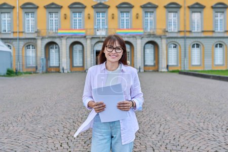 Photo for Outdoor portrait of middle-aged female teacher, mentor, pedagogue posing against background of an educational building, college university - Royalty Free Image