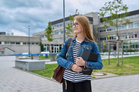 Téléchargez les photos : Jeune adolescente en plein air près du bâtiment de l'école. Femme adolescente souriante avec tablette numérique sac à dos regardant vers le côté, espace de copie. Adolescence, éducation, concept d'apprentissage - en image libre de droit