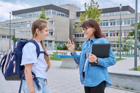 Photo for Talking female teacher and schoolgirl child outdoor, school building background. Meeting communication student girl with backpack and mentor counselor. Education, pre-teenage, learning, back to school - Royalty Free Image