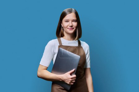 Photo for Portrait of young smiling confident woman in an apron holding laptop in hands, looking at camera on blue studio background. Worker, startup, small business, job, service sector, staff, youth concept - Royalty Free Image