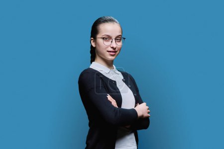 Téléchargez les photos : Portrait adolescent souriant confiant avec des lunettes sur fond bleu. Femme positive 16, 17 ans regardant la caméra avec les bras croisés lycéen style de vie adolescent adolescent - en image libre de droit