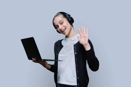 Photo for Teen girl student wearing headphones using laptop, looking at camera on grey studio background. Audio video internet technologies in education learning, online lessons, adolescence high school concept - Royalty Free Image