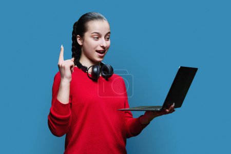 Téléchargez les photos : Portrait d'une adolescente étudiante dans un casque à l'aide d'un ordinateur portable sur fond de studio bleu. Adolescente souriante en rouge regardant l'écran d'ordinateur. Adolescence, éducation loisirs mode de vie technologie - en image libre de droit