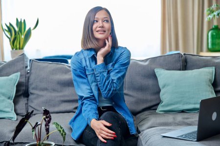 Téléchargez les photos : Portrait de jeune femme attrayante souriante regardant la caméra, assise sur le canapé dans le salon à la maison. Mode de vie, loisirs, concept jeunesse - en image libre de droit
